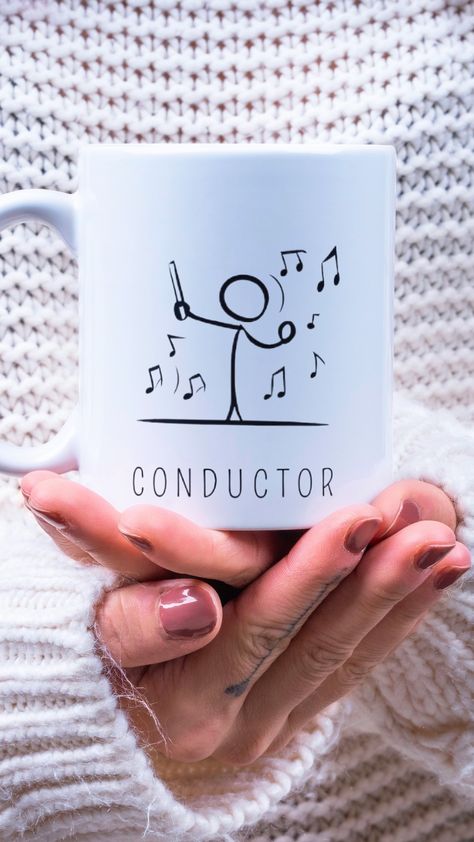 Una taza blanca de cerámica con el diseño minimalista de un director de orquesta (conductor) sobre las manos de una persona.