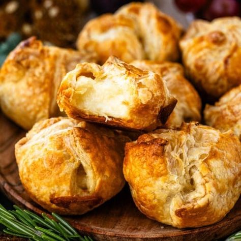 This close-up image highlights a batch of mini baked brie pastries with one pastry cut open to reveal its warm, gooey cheese filling. The golden, flaky crust contrasts beautifully with the soft, melted brie inside, making it look irresistibly delicious. Fresh rosemary sprigs and seeded crackers surround the platter, adding to the rustic and festive presentation. The setup is perfect for a holiday appetizer, inviting guests to enjoy a rich and comforting bite. Mini Baked Brie, Seeded Crackers, Appetizer Thanksgiving, Baked Brie Bites, Melted Brie, Baked Brie Appetizer, Brie Appetizer, Macaron Filling, Fancy Appetizers