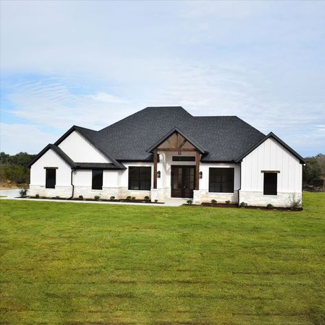 White farmhouse exterior with white vertical siding, black windows, black roof, white rock exterior, cedar trellis and cedar details White Ranch Black Windows Exterior, White Siding Black Roof Farmhouse, White Brick House Farmhouse, White Farmhouse Black Trim Exterior, White House Vertical Siding, White And Black Farmhouse Exterior Ranch, White Brick Black Roof House, White Barndominium With Black Trim, Black And White Modern Farmhouse Exterior With Stone