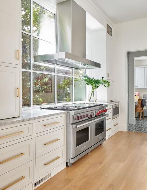 A wolf dual range and hood designed in front of kitchen windows bringing a sheen finish from natural light onto stainless steel. Range Hood In Front Of Window, Cape Kitchen, Kitchen Cooktop, White Shaker Kitchen Cabinets, White Shaker Kitchen, Home Design Magazines, White Shaker Cabinets, Shaker Kitchen Cabinets, White Kitchen Island