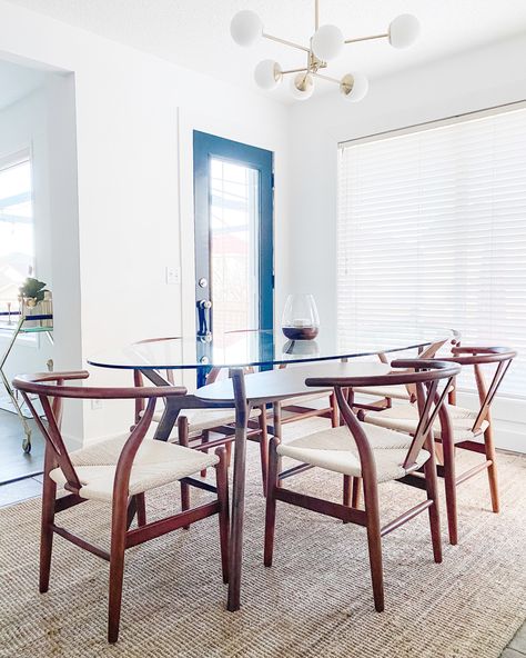 This is a bright dining room with a large window. There is a large jute rug which frames the space, a walnut mid century modern and glass dining table and six walnut wishbone dining chairs. There is a white globe with gold metal, mid century modern chandelier. It’s minimalistic and tidy. The door to the backyard has a large window and is painted black. Glass Table With Wood Chairs, Wishbone Dining Chairs, Glass Dinning Table, Midcentury Modern Dining Room, Modern Glass Dining Table, African Interior Design, African Interior, Wood Chairs, Dining Room Makeover