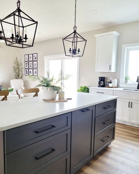 This kitchen features off-white walls and white cabinets with black handles.. Two black pendant lamps hang over a white countertop on a black kitchen island on a warm wood floor. Angular black pulls are attached to the drawers of the island. Black Kitchen Island With Black Hardware, White Kitchen Cabinets Gray Island Black Hardware, Black Cabinet Island, Dark Grey Kitchen Cabinets White Counter, Black Kitchen Island Handles, Black Kitchen Cabinets With Black Pulls, White Kitchen Cupboards With Black Island, White Kitchen Cabinets With Quartz And Black Hardware, Large Black Kitchen Island