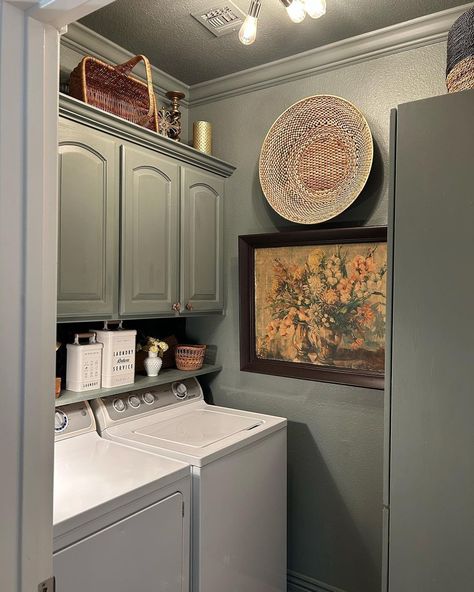 Gray cabinets are mounted above the washing machines in this vintage laundry room, matching the color painted on the walls and ceiling throughout the space. A shelf below the cabinets contains additional storage space, where white tin boxes are displayed. Add a framed painting and a woven wall piece to add character to the space and create an eye-catching design as soon as you enter the room. Laundry Room Painted Ceiling, Color Drenched Laundry Room, Painted Cabinets Laundry Room, Colorful Laundry Room Ideas, Storage Cleaning Supplies, Laundry Room Decorating Ideas, Green Laundry Room, White Cabinets Black Countertops, Rustic Laundry