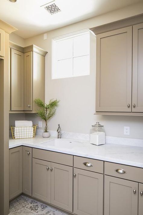 Light brown shaker cabinets accented with satin nickel cup pulls and a white quartz countertop are fitted with a sink matched with a satin nickel faucet located beneath a window flanked by light brown upper cabinets. Brown Painted Cabinets, Light Brown Cabinets, Light Brown Kitchen Cabinets, Tan Kitchen Cabinets, Light Brown Kitchen, Taupe Kitchen Cabinets, Beige Kitchen Cabinets, White Kitchen Countertops, Taupe Kitchen