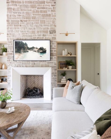 White fireplace with beige herringbone tile firebox is fitted into a floor to ceiling whitewashed stone wall. Black frame beach artwork is mounted above the fireplace in between recessed light wood shelves. Fireplace Sconces, Herringbone Fireplace, Beach Fireplace, White Wash Brick Fireplace, White Wash Brick, Shiplap Fireplace, Fireplace Built Ins, White Fireplace, Wood Fireplace