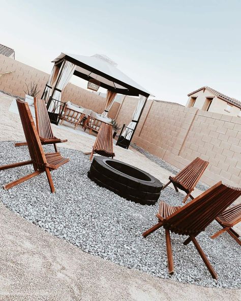 A modern patio has a circle of gray gravel, and a black stone fire pit resting inside. Wooden patio chairs with clean lines are arranged around the fire pit. In the background, a covered pergola is arranged over the top of wooden patio furniture. Wooden Patio Chairs, Wooden Patio Furniture, Wooden Patio, Brick Fire Pit, Fire Pit Ideas, Fire Pit Chairs, Metal Fire Pit, Wood Fire Pit, Stone Fire Pit