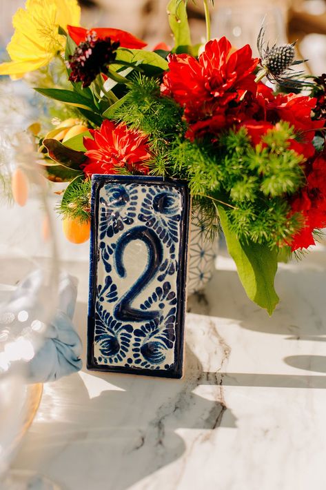 wedding reception courtyard red dahlia thistle greenery blue and white talavera tile table number Mission Style Wedding, Spanish Style Centerpieces, Talavera Wedding Centerpiece, Talavera Quinceanera, Spain Inspired Wedding, Spanish Wedding Table Decor, Spanish Style Wedding Decorations, Talavera Wedding Decor, Spanish Talavera Wedding