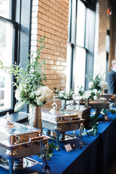 One of the last places couples think to decorate is the buffet table, but sprucing up this space will add some wow factor to your wedding. This couple used a navy linen instead of the typical black, and included some simple vases with a white hydrangea and greenery. Simple, classy, and sure to impress guests. Engagement Buffet Table, Buffet Dishes Display, Chafing Dish Display Ideas Wedding, Buffet Wedding Decor, Rehearsal Dinner Buffet Table Decor, Party Buffet Table Set Up, Buffet Centerpiece Ideas, Elegant Buffet Table Ideas Decor, Wedding Buffet Tables