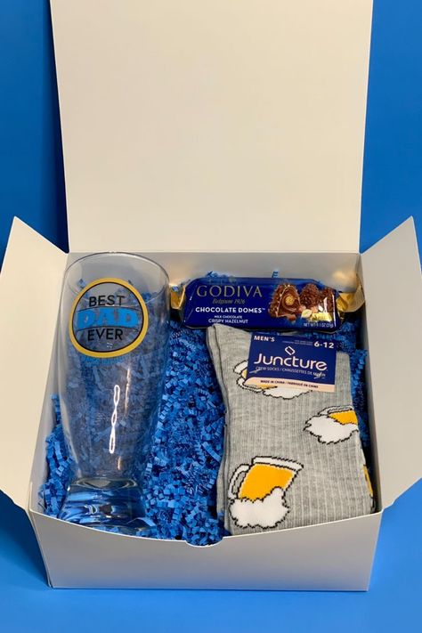 This white paper gift box with blue shred contains: a best dad ever times infinity beer mug with yellow black and blue lettering, a pack of milk chocolate domes with crispy hazelnut, and a grey pair of beer mug crew socks (size 6-12). Beer Gift Basket For Men, Chocolate Domes, Beer Gifts Basket, Gift Box For Him, Chocolate Dome, Fathers Day Gift Basket, Baskets For Men, Gift Baskets For Men, Gift Box For Men