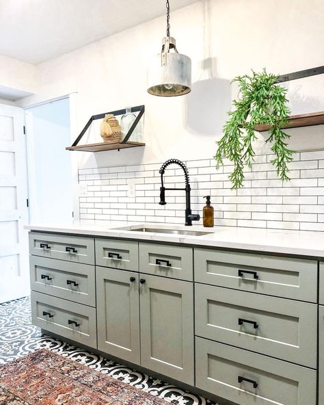 This kitchen boasts white walls, a white subway tile backsplash, a black gooseneck faucet, and a red rug on an ornate black tile floor. Two floating wood shelves hang above the white counter on the light sage green kitchen cabinets with black hardware. Light Sage Green Kitchen, Kitchen Cabinets With Black Hardware, Cabinets With Black Hardware, Sage Green Kitchen Cabinets, Green Kitchen Island, Shiplap Kitchen, Sage Kitchen, Top Kitchen Cabinets, Two Tone Kitchen Cabinets