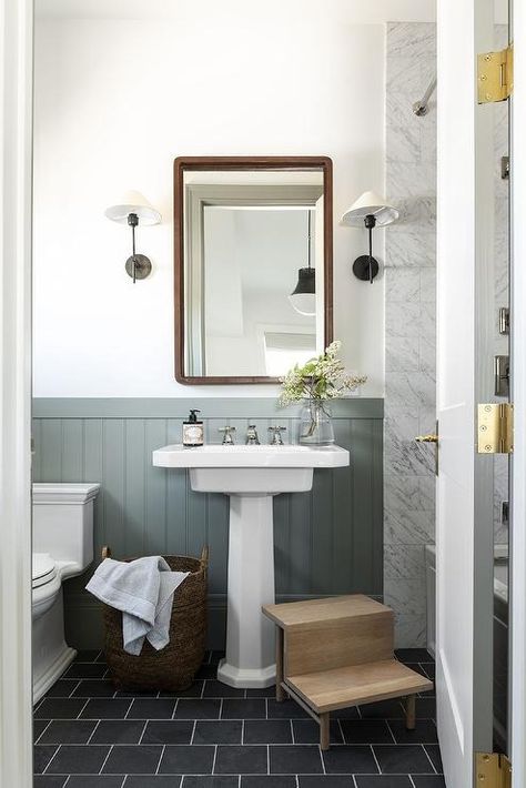 A wooden step stool sits on black brick floor tiles at a white pedestal sink vanity fixed against sage green wall trim beneath a curved brown vintage mirror hung from a white wall between oil rubbed bronze sconces. Pedestal Sink Vanity, Bathroom Pedestal Sink, Black And White Tiles Bathroom, Half Bathroom Decor, Wooden Step Stool, Bronze Sconces, Dark Grey Walls, Wooden Steps, Sage Green Walls