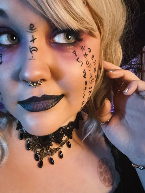 A woman with several runes, glyphs, and magical symbols painted on her face. She has black jewelry, black lipstick, and dark purple eyeshadow above and below her eyes. Necromancer Halloween Costume, Necromancer Costume, Necromancer Outfit, Warlock Cosplay, Dnd Cosplay, Alchemist Cosplay, Magic User, Fair Outfit, Artsy Makeup