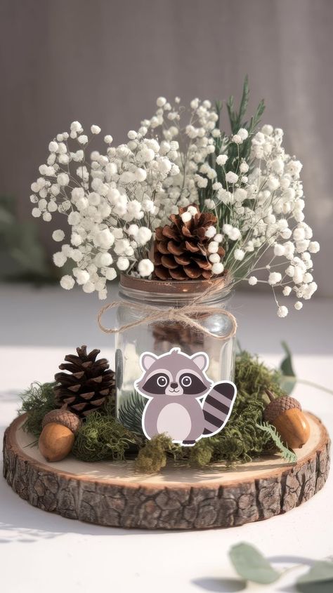 A rustic baby shower centerpiece idea features a glass jar with white baby's breath and pinecones, adorned with a cute raccoon illustration. It sits on a wooden slice surrounded by moss and acorns, creating a cozy, woodland vibe. Forest Baby Shower Theme Centerpieces, Enchanted Forrest Baby Shower Ideas, Baby Boy Centerpiece Ideas, Winter Wonderland Baby Shower Ideas Centerpieces, Nature Baby Shower Ideas, Adventure Themed Baby Shower Ideas, Moose Baby Shower Ideas, Woodland Table Decorations, Outdoorsy Baby Shower Ideas