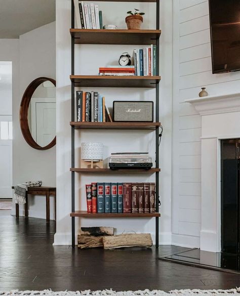 A stained wood and black metal bookshelf is perfect for displaying your favorite books. This shelf is mounted next to a white fireplace mantel fitted to a white shiplap feature wall. A round wood mirror hung above a wooden bench is visible in the background...   Image: decorhint Black Metal Bookshelf, Trending Rugs, White Fireplace Mantel, Chic Bookshelf, Room Bookshelf Ideas, White Fireplace Mantels, Shiplap Feature Wall, White Shelving Unit, Round Wood Mirror