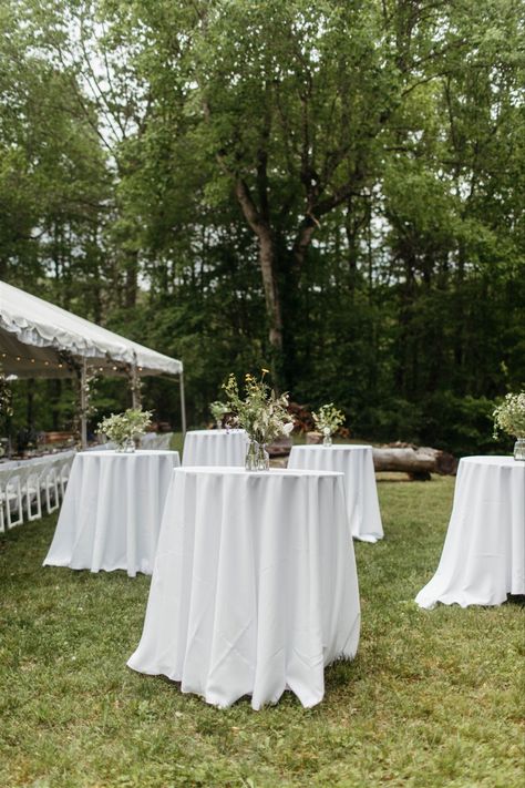 Five cocktail tables with white tablecloths and vases with flowers. Outdoor Wedding Cocktail Tables, Cocktail Hour Layout, Lawn Cocktail Hour Wedding, Simple Cocktail Hour Decor, Cocktail Wedding Layout, Outdoor Wedding Cocktail Hour Decor, Backyard Wedding Cocktail Hour, Easy Backyard Wedding, Outdoor Cocktail Hour Wedding