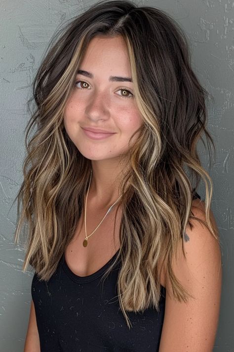 Woman with wavy, highlighted hair smiling against a gray textured wall. Dark Hair With A Few Light Pieces, Brunette Hair Peekaboo Highlights, Brown Fall Hair With Highlights, Brown Hair With Face Highlights, Dark Brown Medium Hair With Highlights, Cute Dark Hair Ideas, Accent Balayage Brunettes, Dramatic Dimensional Hair Color, Brunette Hair With Peekaboo Highlights