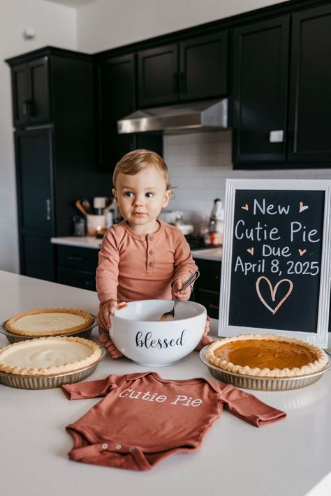 A toddler stands in a kitchen surrounded by freshly baked pies, a charming setup for a November Pregnancy Announcement. A chalkboard sign reading "New Cutie Pie Due April 8, 2025" pairs beautifully with a "blessed" mixing bowl and a onesie labeled "Cutie Pie," crafting a perfect Autumn Pregnancy Announcement. This sweet scene offers a delightful way to reveal a new addition to the family, ideal for Thanksgiving or Fall Baby Announcement ideas. July Pregnancy Announcement Baby 2, Thanksgiving Sibling Announcement, Thanks Giving Pregnancy Announcements, Baby 3 Pregnancy Announcement, Winter Pregnancy Announcement Baby 2, Pregnancy Announcement To Parents Christmas, Fall Baby Announcement Sibling, New Years Pregnancy Announcement Baby 2, Big Brother Announcement Christmas