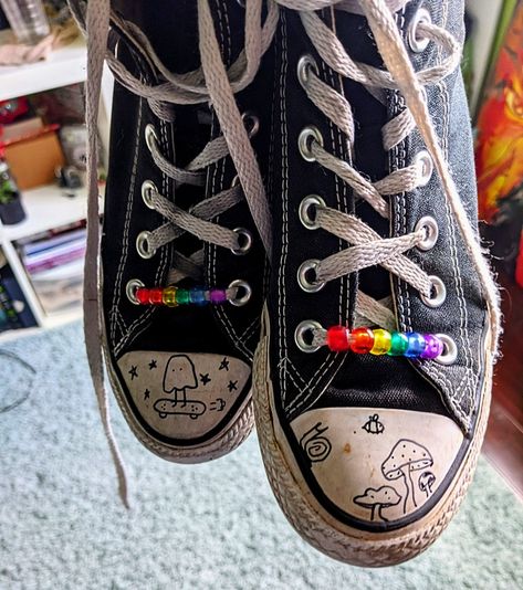 Old black converse all-stars with rainbow beads on the bottom lace. The right shoe has an inked drawing of mushrooms, a snail, and a bee flying. The left shoe has an inked drawing of a ghost on a skateboard surrounded by stars. Converse, Beads, Sneakers, Black