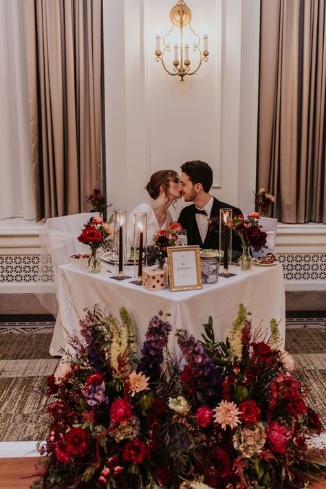 Dark holiday vibes abounded at Celia + Pablo’s “eve of the eve” December 30 wedding. Deep burgundy reds, rich emerald greens, and eggplant purples combined to create a romantic atmosphere. Celia’s bouquet included red garden roses, blue anenomes, purple ranunculus, raspberry stock, and red orchids. Pablo’s boutonniere featured a red orchid with seasonal greenery and berries. Antique gold compotes overflowing with flowers made their  author-themed guest tables gorgeous places to gather. Deep Red And Green Wedding, Deep Red And Purple Wedding, Dark Purple Wedding Theme, Purple And Red Wedding, Head Table Flowers, Moody Wedding Flowers, Purple Ranunculus, Dark Purple Wedding, Red Orchid