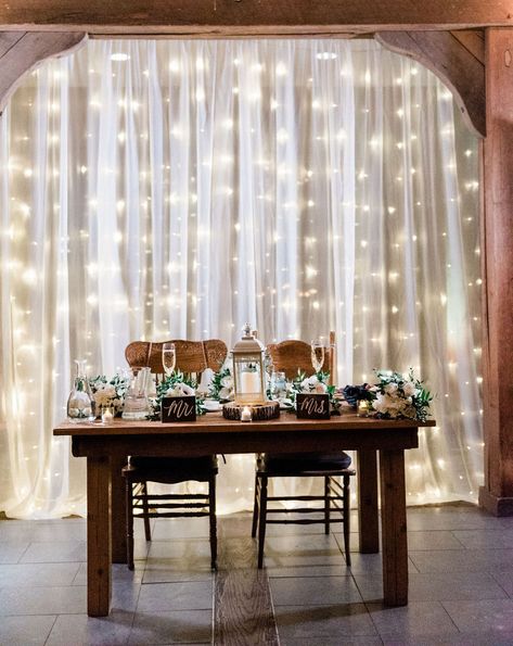 Sheer curtain overlaying lights for a beautiful backdrop behind the sweetheart table Wedding Signs Behind Head Table, Backdrop Curtain Lights, Sweetheart Table Curtain Backdrop, Bride And Groom Table Lights, Curtain Lights Wedding Tent, Curtain Light Backdrop Wedding, Behind Wedding Table Decor, Sheer Curtain Backdrop With Lights, Fairy Light Wall Wedding