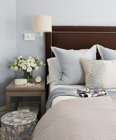 A two tier gray nightstand sits illuminated by a white swing arm sconce mounted on blue walls beside a circular blue floral print stool and a chocolate brown upholstered headboard supporting a bed dressed in a white and black duvet highlighting white shams and blue accent pillows, and a beige, blue and white reef printed lumbar pillow. Brown Headboard, Blue Headboard, Brown Decor, Black Headboard, Transitional Bedroom, Brown Bedroom, Condo Living, Bedroom Black, Trendy Bedroom