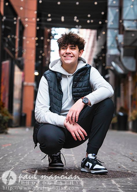 A high school senior boy with curly hair crouches down in an Detroit alleyway decorated with string lights. He is wearing a white hoodie, black puffer vest, black pants, and black and white sneakers. He is smiling and looking away from the camera for his downtown Detroit senior pictures session. The image is by Paul Manoian Photography. Robotics Senior Pictures, Look Down Pose, Boys Poses Photography, Senior Picture Poses For Guys, Chicago Senior Pictures, Golf Poses, Ballet Senior Pictures, Athena Lee, Boy With Curly Hair