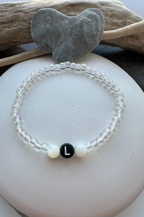 An elastic bracelet with a lack bead with the letter L in white. There are mother of pearl beads either side of the central bead, and clear glass seed beads around the rest of the bracelet. The bracelet is displayed on a white pebble and there are pieces of driftwood and a grey pebble at the top of the photo. Letter L, Elastic Bracelet, Glass Beaded Bracelets, Pearl Gemstone, Initial Letters, Mother Of Pearl, Gemstone Beads, Initials, Glass Beads