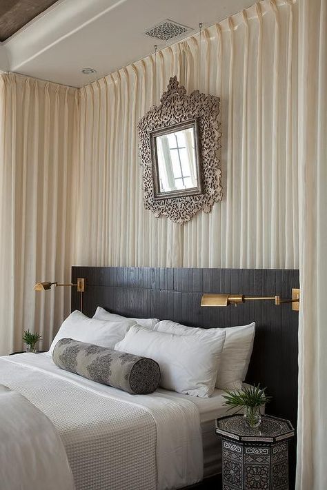 White and black Moroccan style bedroom features walls draped in white sheer drapery panels lined with an ornate mirror placed above a black wood headboard lined with a queen bed dressed in crisp white bedding paired with a black damask bolster pillow as well as a black Moroccan blanket flanked by black octagon shaped accent tables doubling as nightstands illuminated by aged brass swing arm wall sconces, Studio Swing Arm Wall Lights. Light Wood Headboard, Moroccan Style Bedroom, Wall Behind Bed, Curtains Behind Bed, Wall Drapes, Mediterranean Bedroom, Feature Wall Bedroom, Bedroom Drapes, Bedding Inspiration