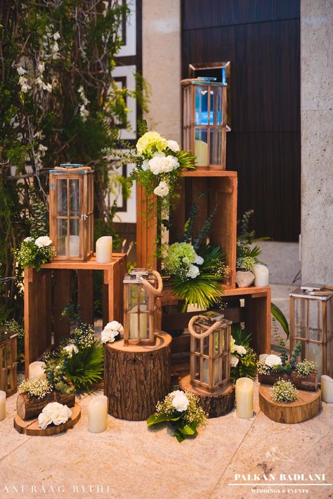 Rustic Decor, Mandap, logs, green, wooden lanterns, white flowers, foilage Green Wooden Wedding, Rustic Wedding Stage Decor, Rustic Stage Decor, Lanterns Stage Decor, Entrance Decor Wedding, Lantern In Trees Wedding, Candle Pathway, Wooden Lanterns Wedding Aisle, Lanterns Hanging From Trees