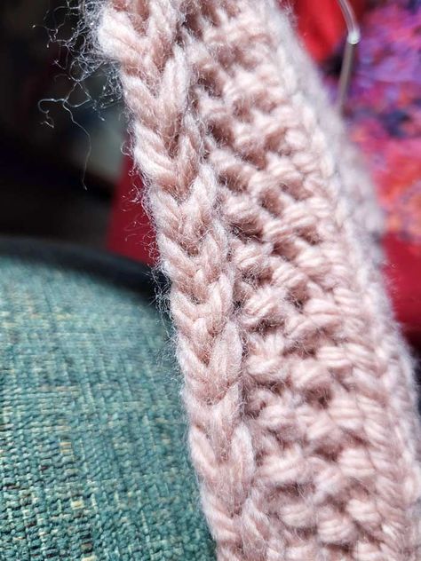 A close-up of the edge of a piece of peach-colored knitting showing a very neat chain edge going up the side of the seed stitch border, with one stitch of the stockinette stitch body of the scarf showing. The background is green on the lower left and red on the remainder; Lion Brand Hue + Me Learn To Knit, Pocket Scarves, Knit Edge, Seed Stitch, Moss Stitch, Lion Brand Yarn, How To Knit, Edge Stitch, Lion Brand