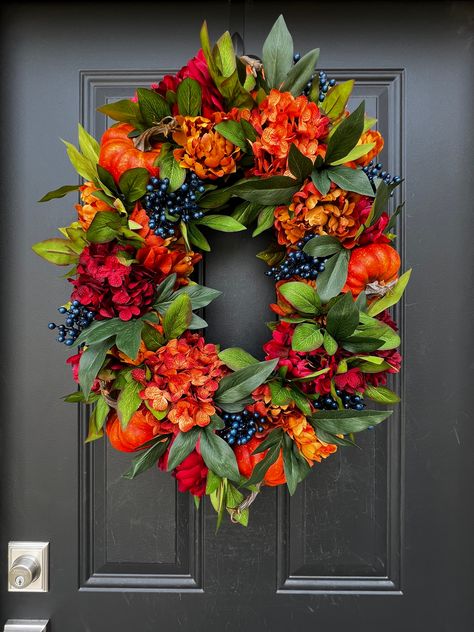 This Oval Bountiful Harvest Wreath brings the warmth and richness of the season straight to your door. Lush, vibrant hues of burnt orange, deep reds, and golden yellows are interwoven with lifelike pumpkins, bold peonies, and clusters of deep blue berries. The oval shape adds an elegant, unexpected twist to the traditional round wreath, making it a striking statement piece. Whether hung on your covered front door or displayed indoors, this handcrafted wreath is perfect for welcoming fall and cel Tombstone Flowers, Centerpiece Craft, Floral Door Wreaths, Blue Berries, Door Wreaths Diy, Harvest Wreath, Bountiful Harvest, Hanging Wreath, Wreath Making