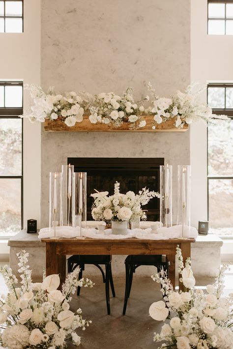 Dramatic white blooms and tall taper candles frame a bridal bouquet on a farmhouse sweetheart table making the perfect table for two. Bridal Bouquet Sweetheart Table, Sweetheart Table Bridal Bouquet, Bridesmaids Bouquets On Sweetheart Table, Sweetheart Table Fireplace, Fireplace Sweetheart Table, Sweetheart Table Wedding Fireplace, Sweet Heart Table Wedding Romantic, Lovers Table Wedding, Fireplace Floral Arrangements