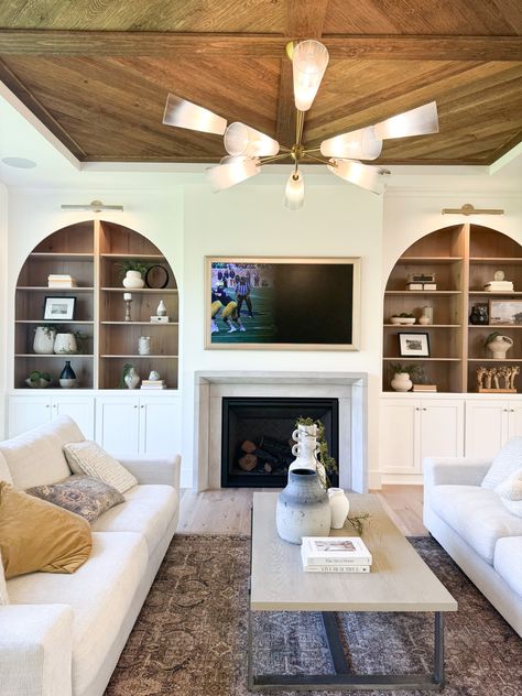 We cannot get over all of the stunning details that were thought of when designing this gorgeous 2024 Fall Parade of Homes living room. The custom arched built-ins that surround the fireplace create an immaculate focal point, with the insets being a rich wood tone that compliment the wood ceiling detail!     New Construction | Home Trends 2024 | Home Design | Minnesota Home Trends Fireplace With Arched Opening, Arched Living Room, Built Ins With Arch, Living Room With Arches, Arched Built In Shelves, Arched Built Ins With Tv, Fireplace With Arched Built Ins, Tv Built Ins, Built In Shelves Living Room Arches