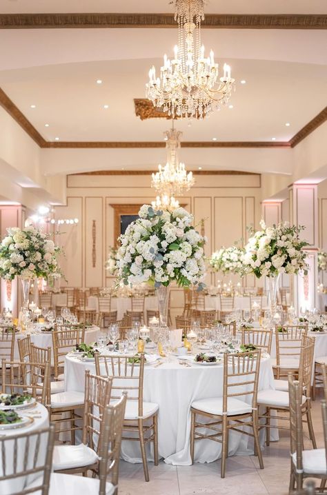 Elegant white ballroom lit by soft lighting from the chandeliers overhead. Tall white floral arrangements adorn the tables and gold chiavari chairs are set at each one. Wedding Reception Ballroom, 5 Tier Cake, Chateau Cocomar, Wedding Chateau, Elegant Wedding Reception, Wedding Venue Inspiration, Wedding Venues Texas, Tier Cake, Ballroom Wedding