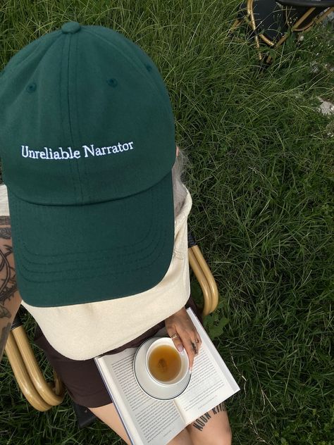 Overhead photo of a green hat that reads 'Unreliable Narrator' worn by a girl with a book and a cup of tea in her lap. She's sitting in a chair in a field. Unreliable Narrator, Good And Bad, Soft Spot, Bad Hair Day, Bad Hair, Baseball Hat, Dad Hat, Hair Day, Trucker Cap