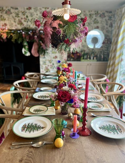 Colourful Christmas tablescape on large wooden table with Spode dinnerware which is white with a green rim and Christmas tree in the centre. Above the table hangs an extravagant foliage cloud filled with greenery, red roses and pink flowers. The centre of the table is fulled with mini floral displays. bright and colourful candlesticks with fruit and baubles scattered throughout. Pink Red Christmas Table, Colourful Candlesticks, Colourful Christmas Table, Spode Dinnerware, Modern Christmas Tablescape, Large Wooden Table, Surviving Winter, Decor Natal, Christmas Table Decorations Centerpiece