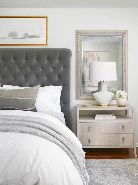 Bedroom features a dark gray velvet tufted sleigh headboard with white and gray bedding, a sliver leaf mirror mounted over a reeded nighstand lit by a white and gold lamp and a gray rug. Grey Velvet Headboard Bedroom Ideas, Grey Tufted Bed Bedroom Ideas, Gray Tufted Headboard Bedroom, Gray Velvet Bedroom Ideas, Grey Tufted Headboard Bedroom, Dark Gray Headboard Bedroom, Dark Grey Headboard Bedroom, Grey Tufted Headboard, Dark Headboard