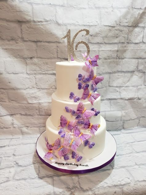 Three tiered Sweet 16 cake in all white fondant with  cascading  butterflies in different shades of purple with 16 rhinestone topper Sweet 16 Cakes With Butterflies, Sweet 15 Butterfly Cake, Sweet 16 Cake Butterfly, Butterfly Themed Birthday Party Sweet 16, Purple And White Sweet 16 Cake, Purple And White Butterfly Cake, Butterfly Party Ideas Sweet 16, Sweet 16 Themes Unique Cake Ideas, Purple Sweet 16 Dessert Table