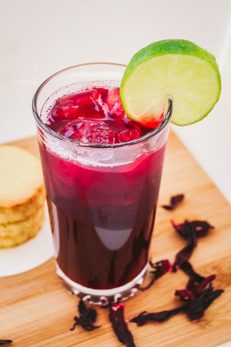 an angled overhead shot of zobo drink garnished with a slice of lime, beside it are stack of cookies Zobo Drink, Hibiscus Drink, Spiced Drinks, Nigerian Recipes, Hibiscus Plant, Sparkling Drinks, Popular Drinks, Drinks Logo, Lime Soda