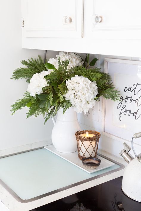 Winter white large head hydrangeas and greenery mixed in a white pitcher becomes cheery winter decor. Christmas Decor With Hydrangeas, Winter White Flower Arrangements, Kitchen Island Flower Arrangement, Winter Floral Arrangements Home, January Flower Arrangements, January Flower, Holiday Floral Arrangements, Winter Floral Arrangements, White Floral Arrangements