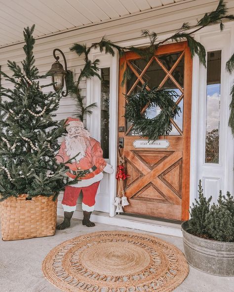 Even Santa will admire this Christmas porch featuring a stained wood farmhouse door with glass. The door is accompanied by a round jute rug, a pine garland, a wreath, and a Christmas tree in a woven basket. Give guests a warm welcome with a vintage Santa standing next to the door. Small Christmas Front Porch Ideas, Front Porch Christmas Decor Ideas Farmhouse, Vintage Christmas Porch, Christmas Welcome Signs, Rustic Farmhouse Front Porches, Decorative Hanging Baskets, Christmas Front Porch Ideas, Round Jute Rug, Black Christmas Decorations