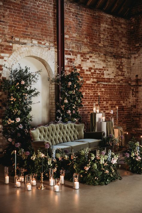 This shows a barn wedding lounge set up. The focus is on a green velvet three seater louis-style sofa. The sofa is surrounded by large flower arrangements and differently sized hurricane lanterns with pillar candles. Next to the sofa are also three ribbed round plinths, two white one wooden. On the plinths are flowers, lanterns and candles. Wedding Portrait Lounge, Photo Lounge Wedding, Portrait Lounge Wedding, Wedding Lounge Seating Indoor, Wedding Photo Area, Lounge Area Wedding, Golden Retriever Wedding, Photo Lounge, Wedding Lounge Seating