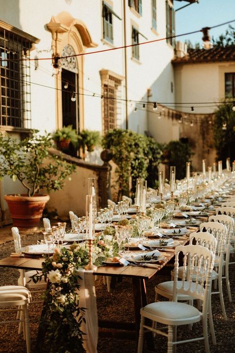 Wedding reception with a beautiful family style long wooden table, white romantic chairs, table runner made of eucalyptus, tea roses and white garden flowers, white taper candles in golden candelholders and vintage table settings under the string lights at a beautiful Italian garden of the 17th century Tuscan villa. White Chairs Wedding, Vintage Table Settings, Italian Dinner Table, White Garden Flowers, Wedding Flower Table Decorations, White Taper Candles, Family Style Weddings, White Flowers Garden, Family Style Table