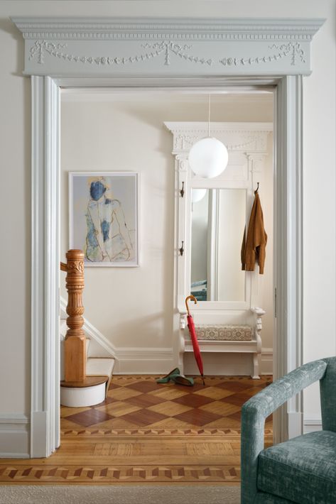 A vestibule featuring traditional decorative molding and trim around the door frame, checkerboard-stained parquet floors, a large Victorian mirror, and a modern blue velvet chair partially visible from the corner, with contrasting colors on the trim and walls. Perfect Fall vibes. Modern art with woman figure. Globe pendant light. Stained Checkerboard Floor, Front Door Vestibule Entryway, Victorian Trim Moldings, Victorian Molding, Victorian Entryway, Blue Velvet Chair, Archways In Homes, Blue Velvet Chairs, Small Foyer