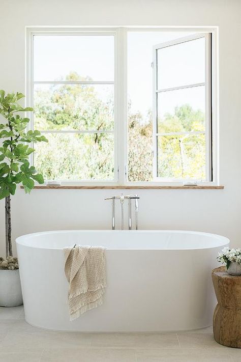 Bathroom features modern oval white bathtub with modern floor mount tub filler under windows, salvaged wood accent table and a potted faux tree. Bath Under Window, Concrete Bathtub, Contemporary Craftsman, Cottage Bathroom, Wood Accent Table, Bathroom Windows, Design Del Prodotto, Bathroom Renos, Shop Interiors