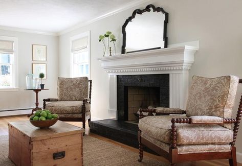 Cottage living room features a white fireplace mantle with dentil moldings accented with a black brick surround and a black brick hearth flanked by a pair of brown spindle chairs upholstered in taupe floral fabric across from a trunk coffee table atop a taupe jute rug. Black Fireplace Mantels, White Fireplace Surround, Black Brick Fireplace, Mantle Design, White Mantle, Brick Living Room, White Mantel, Painted Brick Fireplaces, Black Fireplace