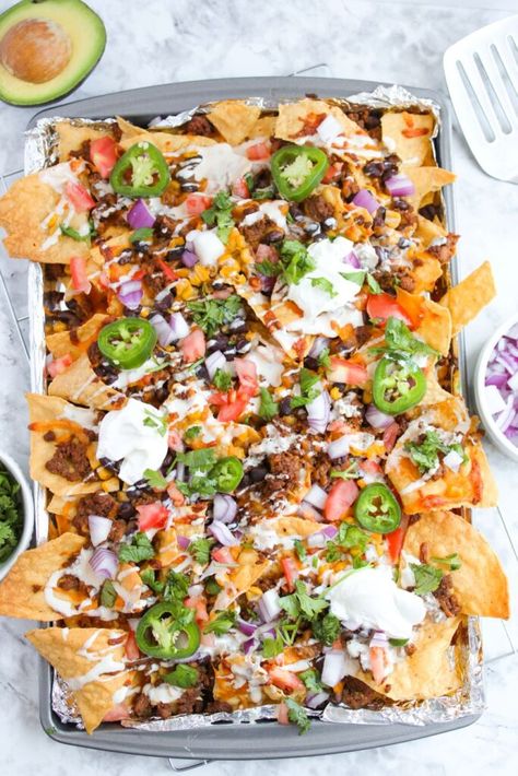 A tray of loaded nachos topped with melted cheese, ground meat, diced tomatoes, red onions, jalapeño slices, cilantro, and dollops of sour cream. An avocado half and a spatula are visible nearby on a marble surface. Nacho Board Ideas, Jalapeno Nachos, Nachos Platter, Nacho Board, Sheet Pan Beef, Sheet Pan Nachos, Pan Nachos, Beef Nachos, Nacho Toppings