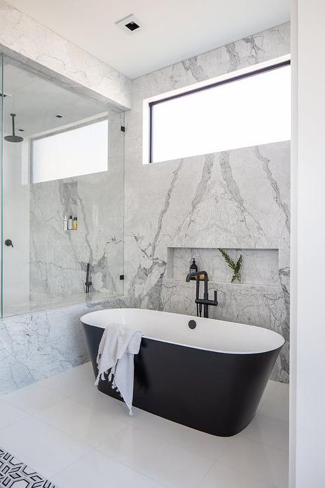 Elegant bathroom boasts a modern black and white oval bathtub with a matte black floor mount tub filler positioned on a white tiled floor beneath a marble tiled niche framed by a bookmatched marble slab wall finished with a picture window. Window Above Bathtub, Tiled Bathtub, Tiled Niche, Shampoo Niche, Tub Bathroom Ideas, Concrete Bathtub, Transitional Bathroom Design, Country Style Bathrooms, Floor Mount Tub Filler