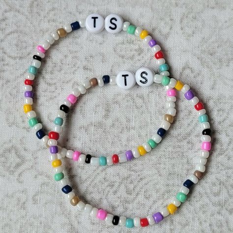 Two Taylor Swift friendship bracelets, made with 3mm seed beads and the letters T and S in the centre. The beads alternate between a colour that represents an era (green, yellow, purple, red, light blue, black, pink, grey, brown and dark blue) and pearl. Eras Tour Seed Bead Bracelets, Taylor Swift Bracelets Seed Beads, Seed Bead Bracelets Taylor Swift, Taylor Swift Seed Bead Bracelet, Era Tour Bracelets Ideas, Swiftie Bracelets, Taylor Bracelets, Eras Tour Friendship Bracelets, Eras Bracelets