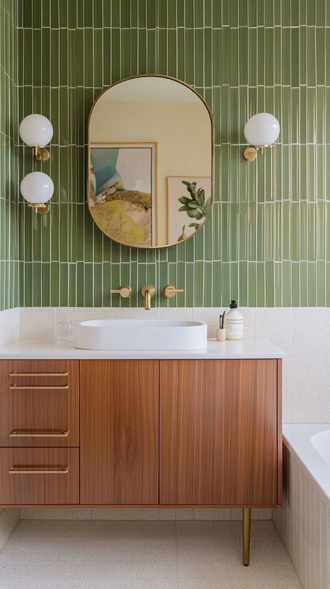 Elegant mid-century modern bathroom with Blu Dot-inspired elements, featuring a freestanding vanity in walnut wood with tapered legs and flat-front doors with brass pull handles, reminiscent of classic mid-century furniture. The countertop is a white quartz slab, paired with a modern, curved brass faucet for a touch of vintage charm. The bathtub area is wrapped in olive green subway tiles with a glossy finish, arranged in a stacked pattern to add a contemporary edge. Above the vanity, an arched mirror with a thick brass frame makes a statement, flanked by retro-inspired sconces with globe bulbs and brass accents. The floor is laid with terrazzo tiles in shades of cream and beige, adding texture and visual interest, while framed vintage artwork on the walls completes the space with a refine Small Bathroom Ideas Mid Century Modern, Mid Century Modern Half Bath, Green And Cream Bathroom, Mid Century Powder Room, Mid Century Modern Bathroom Design, Mid Century Modern Bathroom Vanity, Green Bathrooms Inspiration, Mid Century Bathroom Remodel, Feminine Bathroom